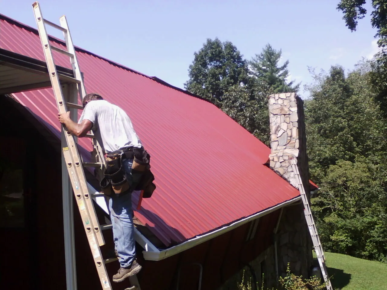 Roofer on a ladder beside a red metal roof and stone chimney — Soffit & Fascia Repair by Roofing Brothers in New Athens